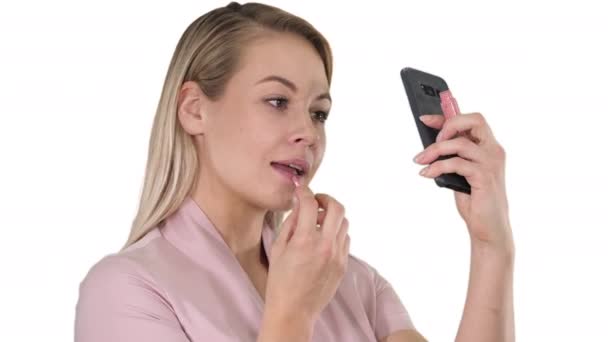 Vacker ung blond kvinna som målar sina läppar med rött läppstift och tittar i telefonen på vit bakgrund. — Stockvideo