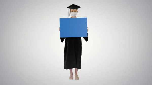 Laureata studentessa in maschera di protezione tenendo bordo vuoto su sfondo gradiente . — Foto Stock