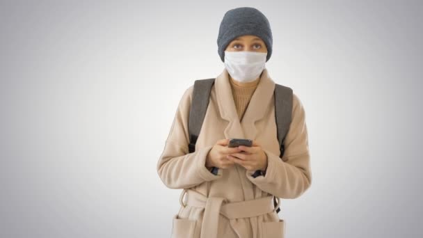 Mujer vestida con un abrigo con máscara médica usando teléfono y caminando sobre fondo degradado . — Vídeos de Stock