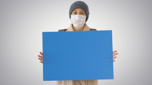 Женщина в наружной одежде и в медицинской маске держит пустую доску на градиентном фоне . — стоковое видео