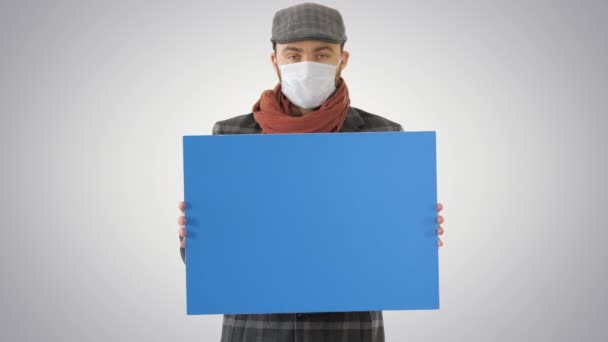 Man i en trendig outfit och medicinsk mask håller tomma plakat på lutning bakgrund. — Stockvideo