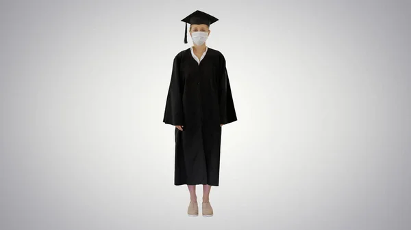 Студентка-випускниця в медичній масці, що стоїть на градієнтному фоні . — стокове фото