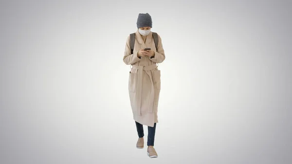 Mulher vestida com um casaco usando máscara médica usando telefone e andando em fundo gradiente . — Fotografia de Stock