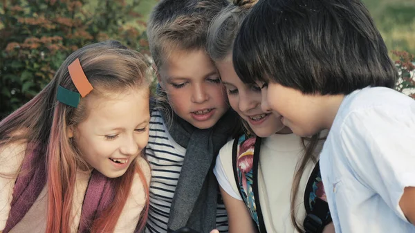 Quatro crianças felizes sentam-se ao ar livre e usam o telefone celular sorrindo . — Fotografia de Stock