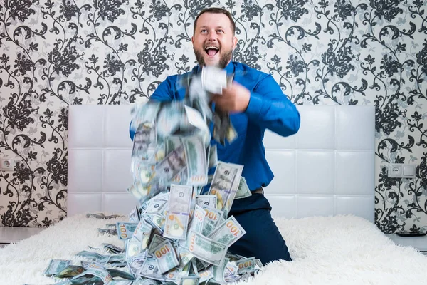 Бородатый мужчина в голубой рубашке наслаждается большими деньгами на белой кровати, много денег летит в воздухе, много банкнот долларов . — стоковое фото