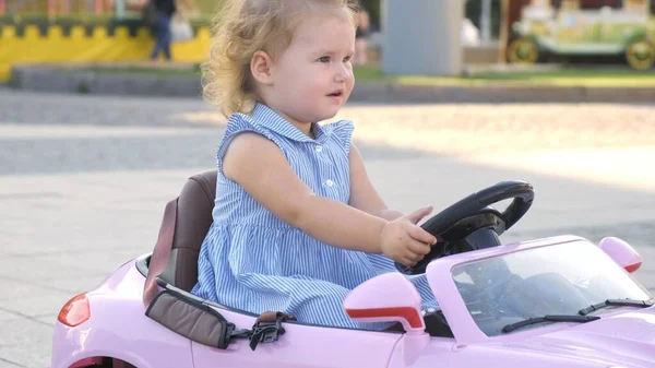 Niña en un vestido azul monta un coche rosa bebé en un parque. Concepto de infancia feliz. Caricatura de una mujer adulta conduciendo un coche . — Foto de Stock