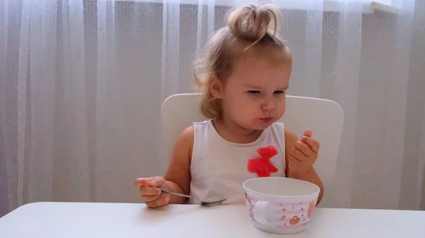 Menina infantil comendo comida de bebê em uma pequena mesa de crianças em um fundo branco. Nutrição e alimentação de crianças . — Fotografia de Stock