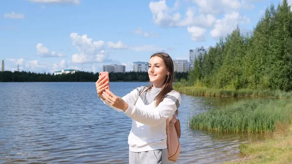 Menina bonita com uma mochila leva uma selfie perto do lago e fala em um telefone celular no Skype . — Fotografia de Stock