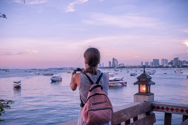 Красивая женщина стоит и фотографирует закат. Счастливая молодая женщина с фотоаппаратом и рюкзаком . — стоковое фото