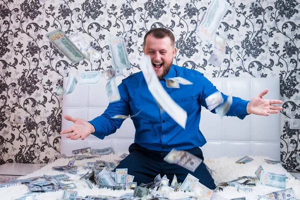 Bearded man in a blue shirt enjoys a lot of money on a white bed, a lot of money flies around a man in the air, a lot of banknotes of dollars — Stock Photo, Image