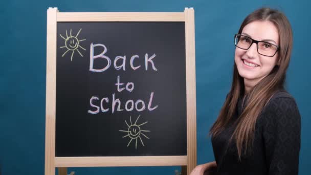 De volta à escola, uma professora com óculos fica perto do quadro-negro da escola e sorri — Vídeo de Stock