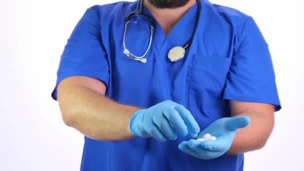 Бородатый врач мужчина в синей одежде и синих резиновых перчатках держит в руках белые таблетки и предлагает вам белый фон, крупным планом — стоковое видео