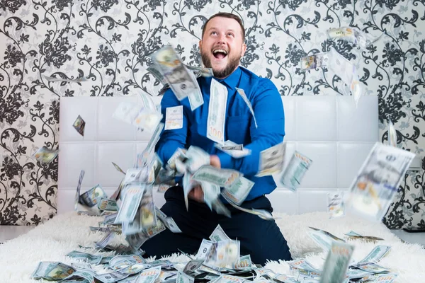 Bearded man in a blue shirt enjoys a lot of money on a white bed, a lot of money flies around a man in the air, a lot of banknotes of dollars — Stock Photo, Image