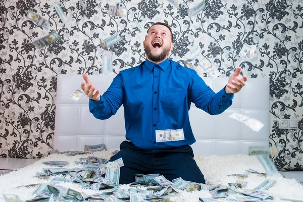 Bearded man in a blue shirt enjoys a lot of money on a white bed, a lot of money flies in the air, a lot of banknotes of dollars. — Stock Photo, Image