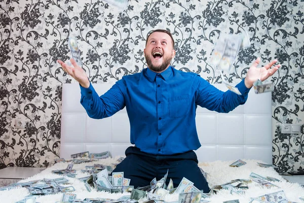 Un uomo in camicia blu gioisce per l'enorme ricchezza sul letto bianco, un sacco di soldi vola in aria . — Foto Stock