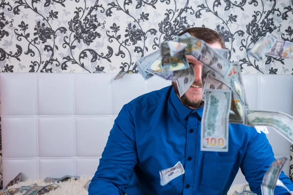 Bearded man in a blue shirt enjoys a lot of money on a white bed, a lot of money flies in the air, a lot of banknotes of dollars. — Stock Photo, Image