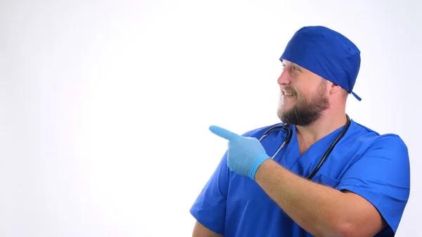 Bärtig lächelnder Arzt in blauer Uniform zeigt seine Hand auf einer leeren Stelle auf weißem Hintergrund. — Stockfoto