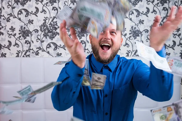 Bearded man in a blue shirt enjoys a lot of money on a white bed, a lot of money flies in the air, a lot of banknotes of dollars. — Stock Photo, Image