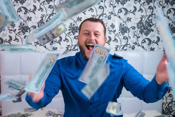 Un uomo in camicia blu gioisce per l'enorme ricchezza sul letto bianco, un sacco di soldi vola in aria . — Foto Stock