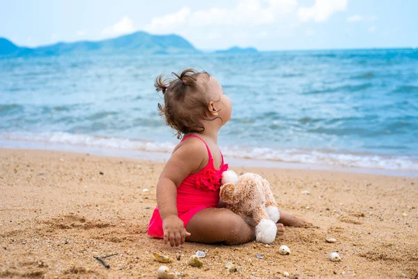 Задумчивая маленькая девочка обнимает плюшевого мишку и смотрит в сторону, сидя на берегу моря . — стоковое фото