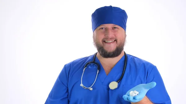 Γενειοφόρος γιατρός άνθρωπος σε μπλε ρούχα και μπλε γάντια καουτσούκ κρατά λευκά χάπια στα χέρια του και σας προσφέρει ένα λευκό φόντο, κοντινό πλάνο — Φωτογραφία Αρχείου