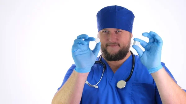 Barbudo médico homem em roupas azuis e luvas de borracha azul segura pílulas brancas em suas mãos sobre fundo branco, close-up — Fotografia de Stock