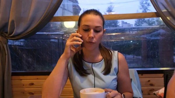 Žena v modrých šatech pije kávu a večer mluví po telefonu v kavárně — Stock fotografie