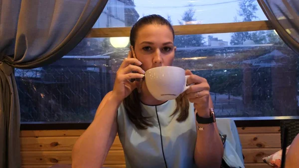 Feminino em um vestido azul bebe café e falando ao telefone em um café à noite — Fotografia de Stock