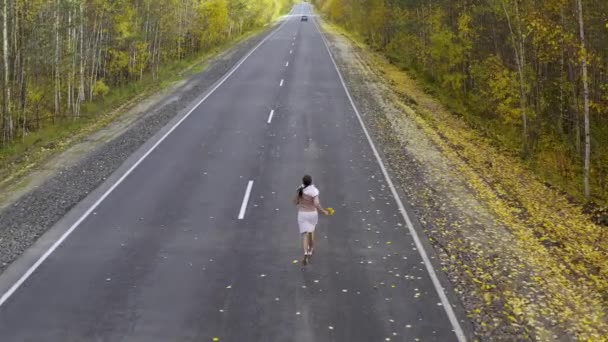 Eine Frau in pinkfarbener Kleidung läuft und wirbelt auf einer asphaltierten Straße inmitten eines herbstlichen Waldes. Ansicht von oben — Stockvideo
