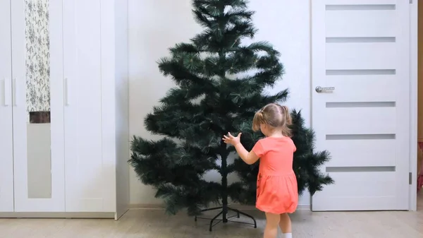 Niña está de pie con su espalda a la cámara y admira el árbol de Navidad. Una niña de 2 años ve un árbol de Navidad por primera vez y se regocija. Inmersión en un cuento de hadas . — Foto de Stock