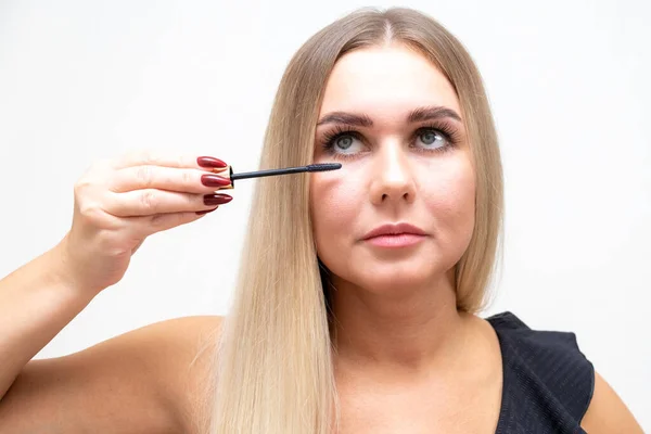 Vrouw zet zwarte mascara op haar wimpers met make-up, foto van aantrekkelijke blonde op een witte en grijze achtergrond. — Stockfoto
