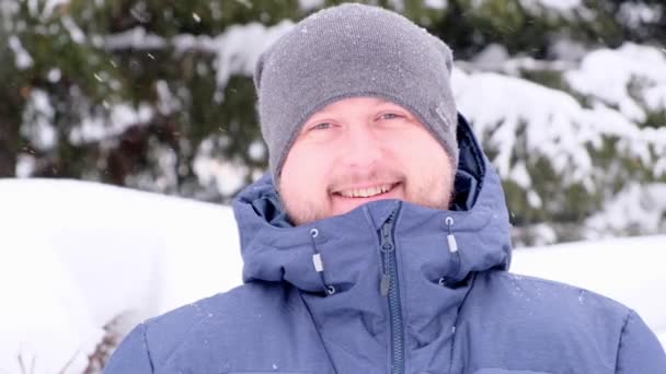 Macho com uma barba em roupas azuis de inverno gosta de cair neve em um dia ensolarado de inverno, neve caindo. Retrato de um homem sorridente no inverno — Vídeo de Stock