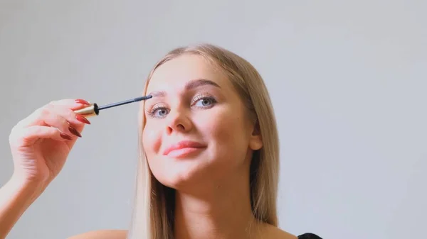 Woman puts black mascara on her eyelashes with makeup, photo of attractive blonde on a white and gray background. — 图库照片