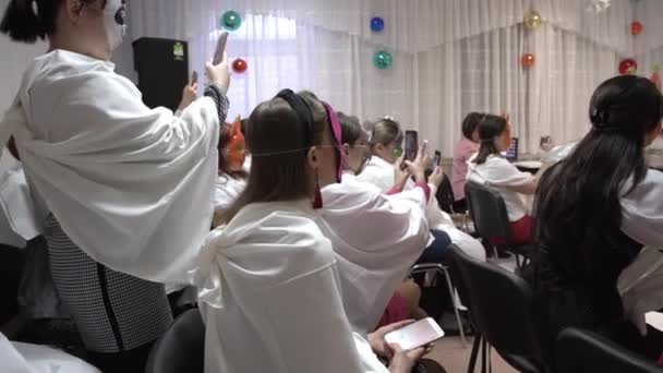 Nizhnevartovsk, Rusia - 12 de diciembre de 2019: Las personas enmascaradas en sus rostros se sientan en sillones y observan el desempeño de sus hijos. La gente está filmando un teatro en un smartphone . — Vídeos de Stock