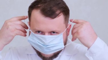 Sakallı erkek doktor koruyucu bir maske takmaya çalışıyor. Hijyen konsepti. Mikrop ve bakterilerin yayılmasını önler ve taç virüsü bulaşmasını önler.
