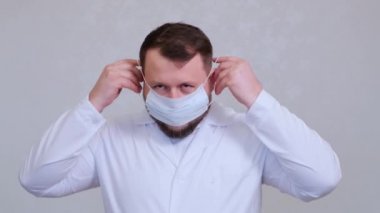 Sakallı erkek doktor koruyucu bir maske takmaya çalışıyor. Hijyen konsepti. Mikrop ve bakterilerin yayılmasını önler ve taç virüsü bulaşmasını önler.