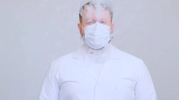 Macho em roupas brancas fuma um cigarro eletrônico através de uma máscara médica protetora. Proteção contra vírus, coronavírus, doenças pulmonares — Fotografia de Stock