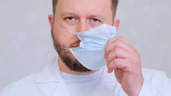 A szakállas férfi orvos szomorú tekintettel veszi le védőmaszkját. Ez a vég. Higiéniai koncepció. megakadályozza a baktériumok és baktériumok terjedését, és elkerüli a koronavírussal való fertőződést — Stock Fotó
