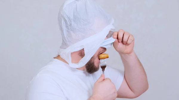 Bearded man in a bag on his head and a protective mask has a cutlet from a fork. Fear of infection. Psychological assistance in case of panic. Joke situation — Stock Photo, Image