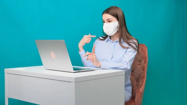 Eine Frau mit Schutzmaske spricht per Videotelefonie, die Finger auf einem Laptop am Arbeitsplatz oder zu Hause während einer Pandemie zeigt. Das Konzept der Arbeit während Quarantäne und Selbstisolierung. — Stockfoto