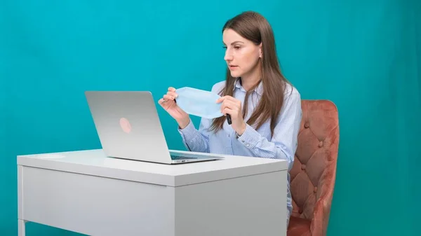 La donna indossa una maschera protettiva mentre lavora su un computer portatile sul posto di lavoro o a casa durante una pandemia. Il concetto di lavoro durante la quarantena e l'autoisolamento . — Foto Stock