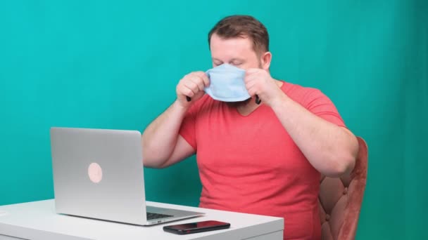 Um homem barbudo em uma camiseta rosa coloca uma máscara médica protetora para trabalhar em um laptop em casa em um fundo verde. Conceito de quarentena — Vídeo de Stock