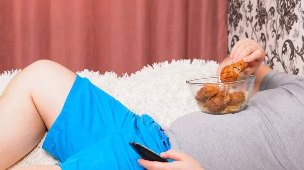 Hombre gordo con un vientre grande está acostado en el sofá con comida rápida y se aburre delante de la televisión con el control remoto en su mano, primer plano . — Foto de Stock