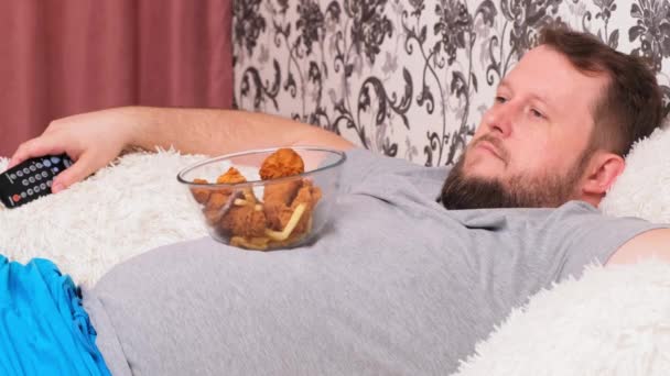 Koca göbekli şişman bir adam elinde fast food ile kanepede yatıyor ve elinde uzaktan kumandayla televizyonun önünde sıkılıyor.. — Stok video