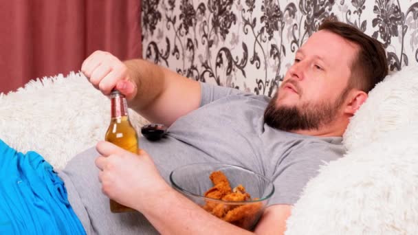 Barbudo gordo con un gran vientre se encuentra en el sofá con comida rápida, abre una cerveza y se pierde el televisor con el control remoto en la mano, de cerca . — Vídeos de Stock