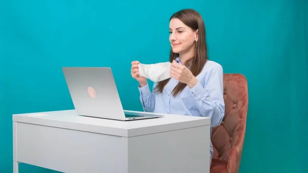Femeia pune o mască de protecție în timp ce lucrează la un laptop la locul de muncă sau acasă în timpul unei pandemii. Conceptul de muncă în timpul carantinei și auto-izolării . — Fotografie, imagine de stoc