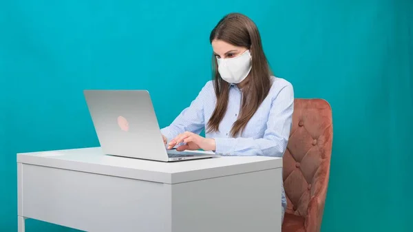 Die Frau mit Schutzmaske arbeitet während einer Pandemie am Laptop am Arbeitsplatz oder zu Hause. Das Konzept der Arbeit während Quarantäne und Selbstisolierung — Stockfoto