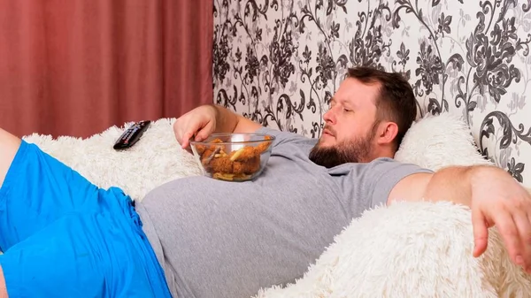 Жирний чоловік з великим животом лежить на дивані з фаст-фудом і нудно перед телевізором з пультом в руці, крупним планом . — стокове фото