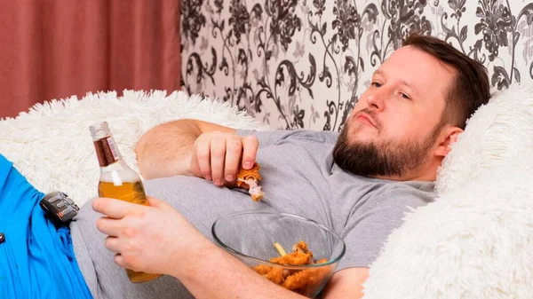 Barbudo gordo con un gran vientre se encuentra en el sofá con comida rápida, bebe cerveza y echa de menos el televisor con el mando a distancia en la mano, primer plano . —  Fotos de Stock