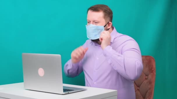 Een man met een baard in een lila shirt doet een beschermend medisch masker af voor het thuis werken op een laptop op een groene achtergrond. Quarantaine-eindconcept — Stockvideo